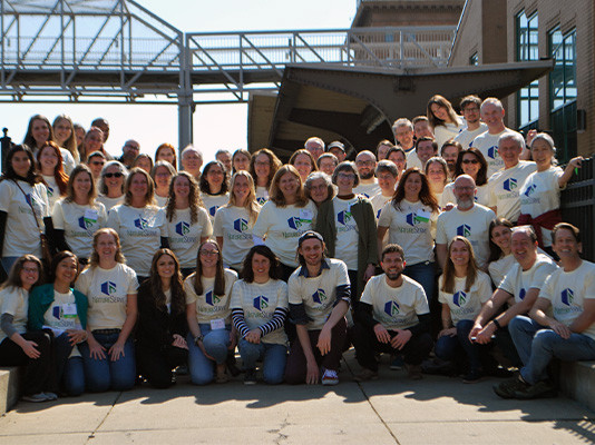 A large group of people wearing shirts with NatureServe's logo are posing under a bridge.