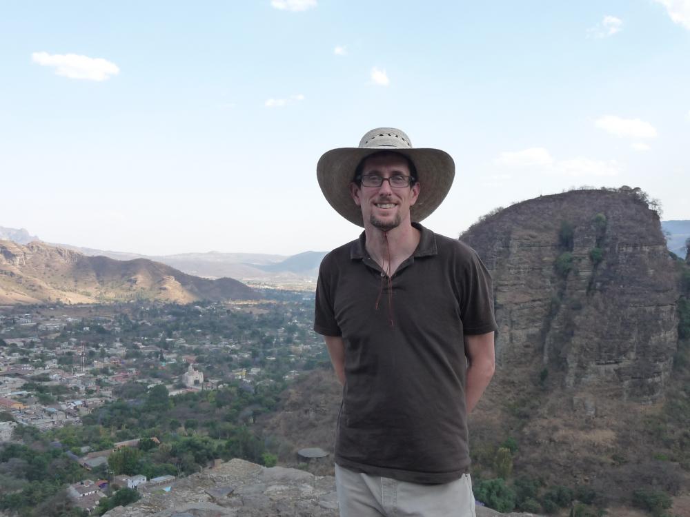 Patrick McIntyre stands on a vista, with blue sky, a valley, and mountains in the background. He wears a wide-brim hat and a brown short-sleeve collared shirt.